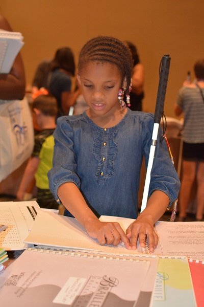 A young girl with a white cane reads from a Braille book.