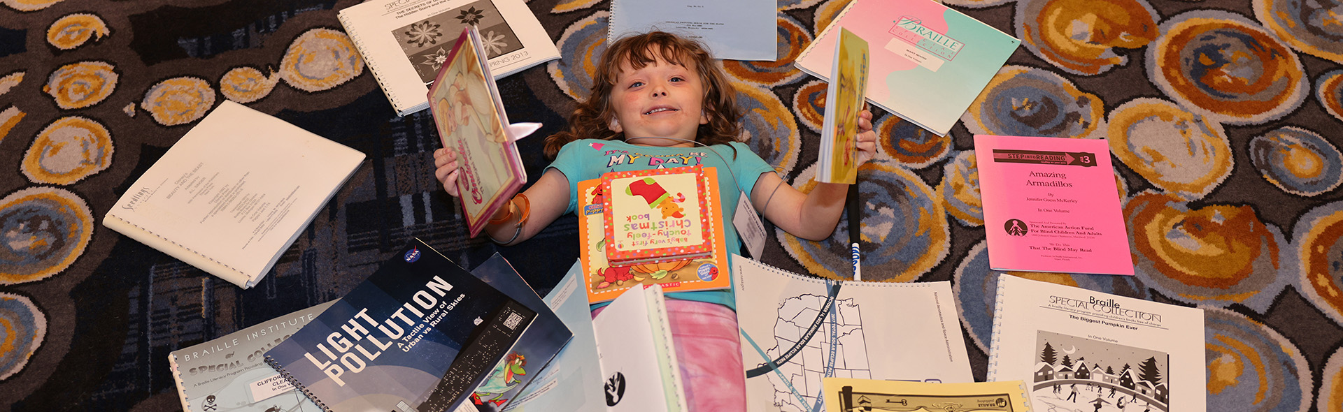 A young blind girl smiles while lying on the floor under a pile of Braille and Twin Vision books.